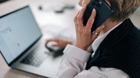 Getty Images A woman with dark blonde hair cut into a short pixie cut. She is wearing a white shirt underneath a black pinstripe vest. She is holding up a phone to her ear while she sits at a desk with her laptop open.