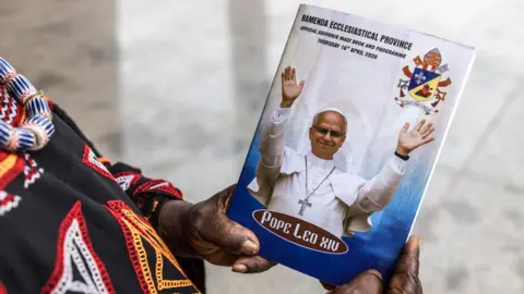 A Catholic worshipper at the Saint Joseph Catherdral in Bamenda, holding a Mass book with the image of the Pope on it. Only the worshippers hands and partial clothing is seen.