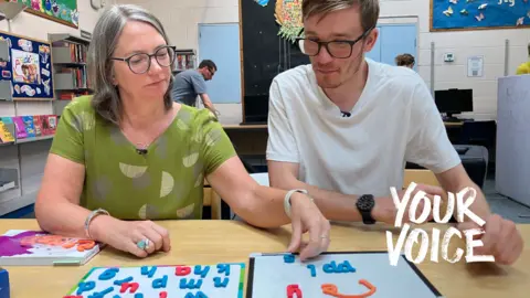 Two people sit at a table in a library, using magnetic letters on whiteboards as part of a literacy or spelling activity. One person places a blue letter on a board while the other watches. Colourful alphabet pieces are spread across the table, with bookshelves, computers, and children's artwork visible in the background.