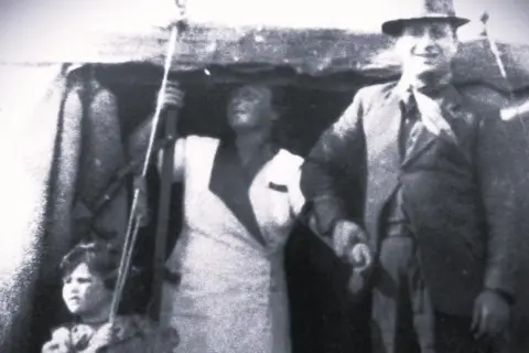 Josephine Smith A man wearing a suit standing next to a woman in a white outfit and a child as they set up a tent.