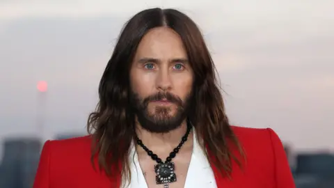 Getty Images Jared Leto wears a red jacket over a white shirt. There is a large black necklace around his neck. 