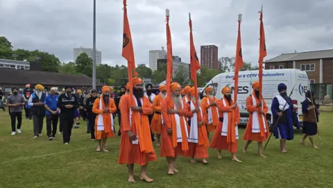 A group of people take part in a procession across a green lawn. The two front rows are made up of men wearing orange clothes and orange turbans. The five men at the very front are each holding an orange flag.