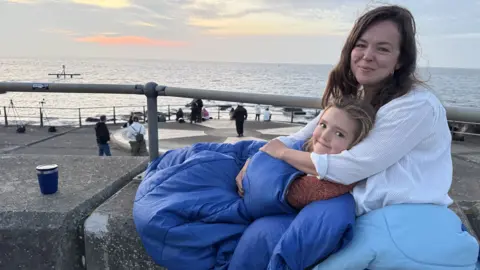 Zoie O'Brien/BBC A woman and young girl smiling as they sit on a concrete sea defence wall, the woman cuddling the girl from behind as she reclines back against her chest. The woman has brown hair and a white top, the girl is blonde and wrapped in a blue sleeping bag. Behind them is a grey sea and the horizon 