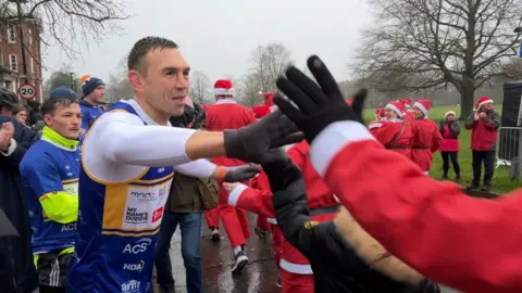 BBC/Michelle Lyons Kevin Sinfield, wearing a blue and yellow athletic vest with charity logos, is giving high-fives to participants dressed in Santa Claus costumes during an outdoor festive event