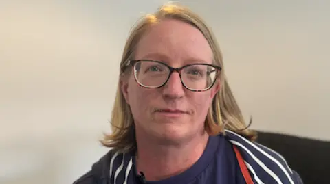 BBC A woman with shoulder-length blonde hair, glasses and wearing blue and white striped top, sitting on a sofa