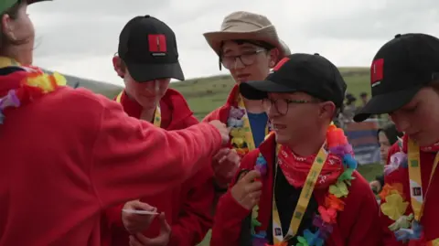 Alfie stood with his team who are wearing red fleeces and Hawaiian flowe necklaces, along with their medals which have yellow straps. They are wearing black baseball caps, and one boy is wearing a cowboy hat. 