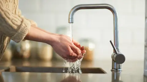 A silver tap running water with someone's hands cupped underneath it. They are wearing a woolly beige jumper and the background is blurred.