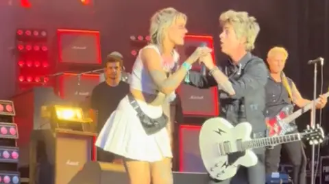 India Barton sharing the mic with Billie Joe Armstrong. He is in all black with a white guitar. She is in a white cheerleader outfit.