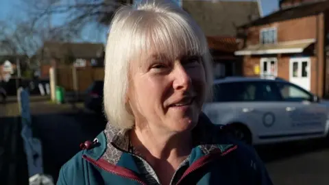 BBC/Qays Najm Hazel Hallett with short blonde hair and wearing a green and maroon winter coat is smiling in the sunshine. She is standing in a town car park and shops and a car can be seen in the background