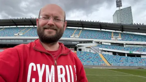 Ian Johnson A man taking a selfie in the Ullevi Stadium in Gothenburg. The stands are empty and he is to the left of the picture. He is smiling at the camera, he has a bald head and has a dark brown beard and moustache. He is wearing rimless glasses and has a red jumper on with the words Cymru emblazoned in white.