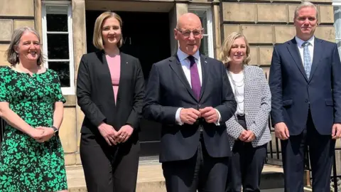 Five people pictured on the sandstone steps of a building. One lady with grey hair in a middle parting is wearing a black and green dress. Next to her is a blonde lady wearing a pink top and a black trouser suit. A bald man in a dark grey suit, white shirt and purple tie is in the middle. To his left is a blonde lady with a white top, black trousers and a black and white patterned jacket. To her left is a man in a navy suit with a white shirt and blue patterned tie. He has grey hair combed in a side parting. 