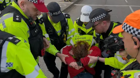 PA Media A group of police officers gather around a woman and grab onto her. She appears to be squatting on the floor and we can only see the top of her head, not her face. The officers wear high-vis vests, black gilets and caps with the word "Police" written on them.