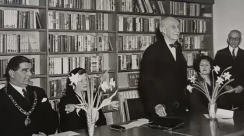 Friends of Woodston Library Five people, in a room, in a black and white image. Two men are standing up and three people are sitting down, in front of shelves of books. The are two floral arrangements on a table. 