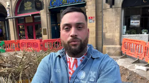 A man with a short brown bears and hair, wearing a white and red T shirt and a blue denim overshirt