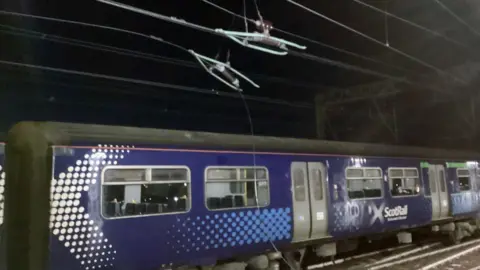 Network Rail A train with blue ScotRail branding underneath a damaged overhead line, drooping down from wires above. 