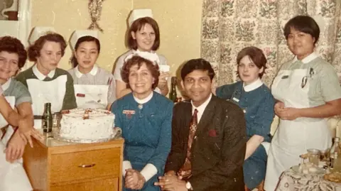 Rotherham NHS Foundation Trust Dr Mondal sits with seven women in nursed uniforms around him. There is a cake on the table. It looks to be the 70s or 80s.