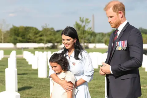 Lindsay Hart for The Office of The Duke of Sussex Prince Harry stands in a suit with military medals next to a mother who has one arm around her young daughter. They are standing on a green graveyard honouring military personnel with white gravestones.