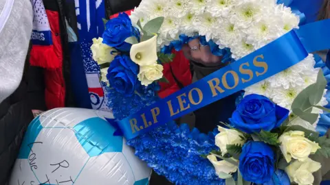 A wreath which reads RIP LEO ROSS. A ringed finger is holding it. Football scarves can be seen in the background. A balloon is also cropped.
