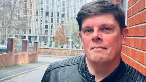 Family photograph Dr Dean Kirby leans against a brick wall in Angel Meadow. He has black short hair and is wearing a black leather jacket. There is a block of flats in the background.