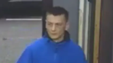 Police Scotland A CCTV image of a man walking into a newsagent wearing a bright blue jacket. He has short dark hair.