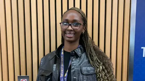Siyon is smiling at the camera. She is wearing a black t-shirt with a black crocodile leather jacket and small silver hoop earrings. There is a blue and yellow university lanyard around her neck. She has black glasses and long blonde braids. Behind her is wood panelling.