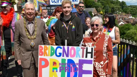 Huw has short blonde hair and is wearing a black hoodie. He jointly holds a Bridgnorth Pride poster, along with mayor David Cooper and mayoress Nicky Cooper.