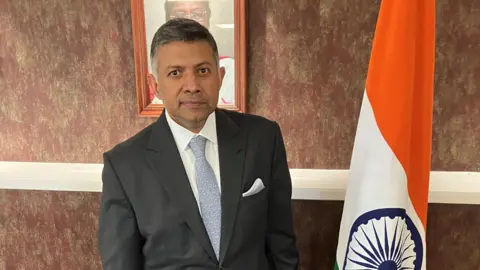 BBC Vikram Doraiswami, High Commissioner of India to the United Kingdom. He is wearing a grey suit and standing next to an Indian flag.