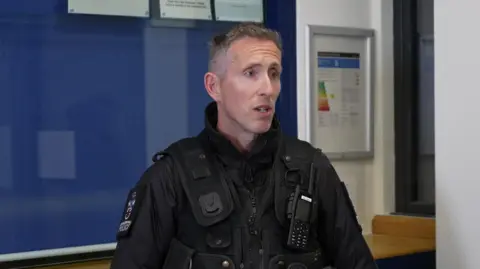 A police officer in police uniform. He is looking to the right. There is a blue and white wall behind him. 