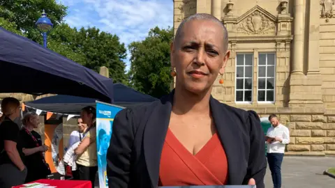 Woman stands in front of a stall, with a stone building in the background, wearing a blazer and red dress, looking at the camera 