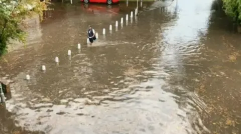 A person wearing shorts and a dark blue gilet, strides through floodwater, from which a line of bollards sticks up