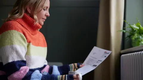 Getty Images Woman in thick jumper sits by a radiator holding a mug and an energy bill.