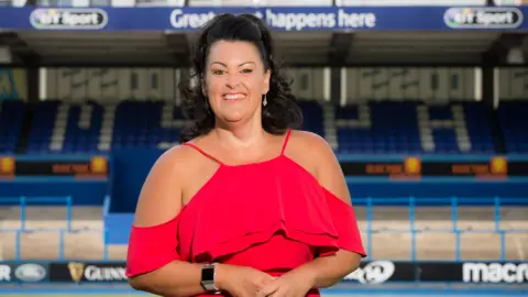 Huw Evans Picture Agency Hayley Parsons smiles at the camera in stood in the Cardiff Rugby stadium. She wears a red dress and drop earrings and her long dark hair is tied back. Blue seats can be seen in the stand behind her. 