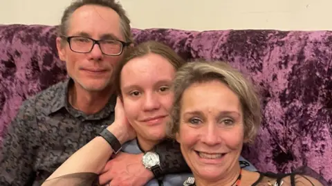 Family photograph Emanuel, Wiktoria and Claudia all sitting on a burgundy velvet sofa hugging one another. They are all smiling. 