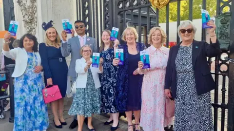 Sbba Siddique Seven women and one man, all smartly dressed in dresses or suits, standing in front of the black gates of Buckingham Palace. They're smiling and holding up leaflets for cancer charity Target Ovarian Cancer.
