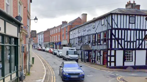 A Google Street View image of Bromyard High Street. There is a blue BMW car driving along a narrow street. There's a row of stationary cars are parked on the right hand side of the road behind it, in front of some local shops and the Tudor-style Kings Arms pub. 