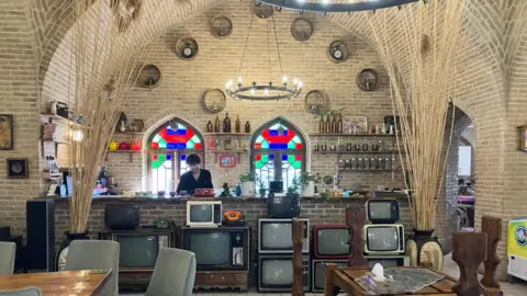 A roadside restaurant on the highway to Tehran. A waiter stands behind a bar making drinks. Lots of old TV screens decorate the wall underneath the bar top.