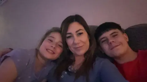 EMAS Sian Deeks sat on a sofa with long brown hair smiling with two children. A girl is sat on her left leaning in and smiling and a boy with black short hair and a red top is also leaning in and smiling.