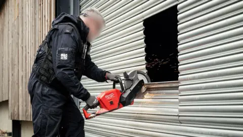 Lincolnshire Police A person whose face is blurred out and has short grey hair is using a red industrial saw to break open a metal wall. A black hole can be seen in a square shape where he is cutting, with sparks coming off the saw. He is wearing all black.