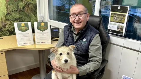 HARPA Skye, a white and brown mixed terrier. She is say on her owner's lap inside an office, with awards for exceptional service on show on a desk next to them. Owner Ray Smith is sat on an office chair. He has short grey hair and blue eyes. He wears glasses, a green shirt and a blue puffer gilet on top.