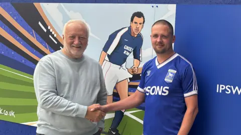 Ipswich Central Steve Flory and Bradley Lloyd standing in front of a mural opposite Portman Road Stadium. Both men are shaking hands and are looking directly at the camera and smiling. Steve Flory on the left is wearing a grey sweatshirt and Bradley Lloyd is wearing a blue Ipswich Town FC shirt. 