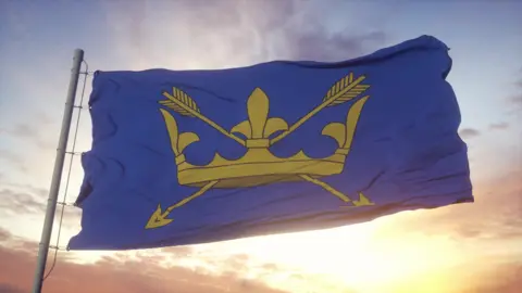 The Suffolk flag is flying in the sunset, taking up most of the picture. The flag has a blue background with a golden crown pierced by two golden arrows. It represents St Edmund, the county's patron saint and last King of East Anglia. 