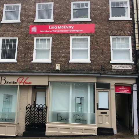 A brick building on Darlington's Blackwellgate. The ground floor has glass windows and signs for a beauty shop. On the right-hand side is an entrance with a small red sign above the doorway for Lola McEvoy's office. A large red banner is attached to the front of the building above the second floor with Lola McEvoy's name on it in white writing. Underneath it says Member of Parliament for Darlington.