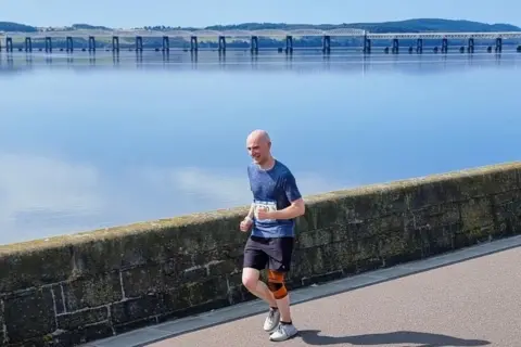 Thomas Mills Thomas Mills runs along the still River Tay wearing a blue top, black shorts, white trainers and an orange knee brace. He smiles at the camera.