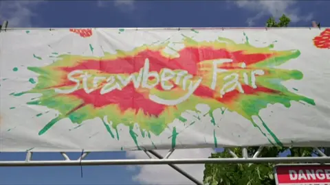 A sign is above an entrance to a festival. The white sign has green, red and yellow paint splashed in the middle which has Strawberry Fair written in white in the middle.
