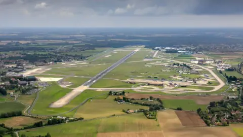 Getty Images An aerial photograph of RAF Lakenheath on a sunny and cloudy day. A long runway can be seen in the middle of the site with taxiways that extend off of that runway. Other airbases buildings can be seen to the left of the image. Green fields surround the airbase.