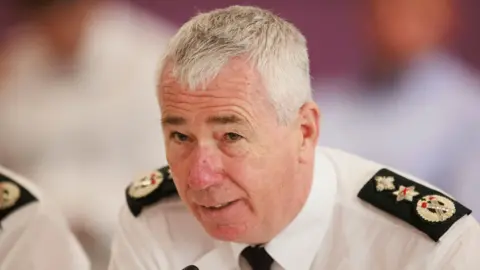 PA Media PSNI Chief Constable Jon Boutcher speaking during a meeting of the Northern Ireland Policing Board in Belfast. He has white hair and wears a white shirt and police shoulder lapels. 