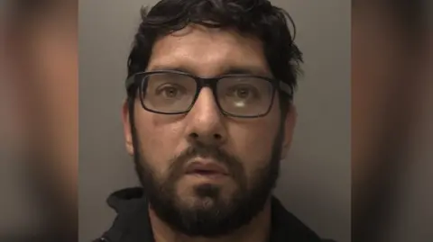Abdul Kahar pictured against a grey wall in a mugshot. He has messy black hair and wears square framed black glasses. He has a black short beard and wears a black zip up hoodie over a round neck blue shirt.