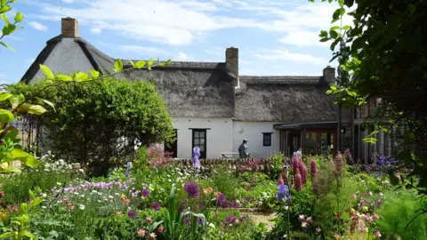 John Clare Cottage A white cottage with a thatched roof is behind a fence in the background. In front is a small garden with different types of flowers and a small tree which is on the left. 