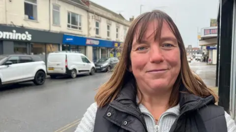 Andrew Turner/BBC Emma Washbourne has long auburn hair and is wearing a grey jersey with a black padded gilet over the top. She is standing on Gorleston High Street, with parked cars and vans in the background, lined up in front of a parade of shops