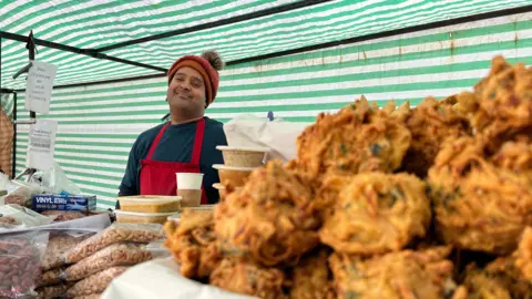 Imran smiles for the camera from behind his counter. Some sort of baked food is in the foreground next to some bags of nuts.